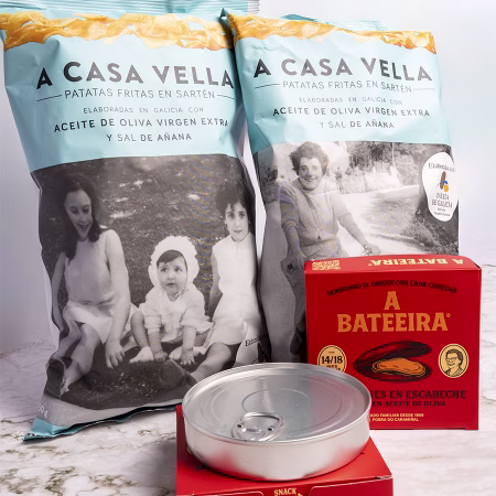 Bolsas de patatas fritas A Casa Vella junto a caja y lata de mejillón, con foto familiar de fondo